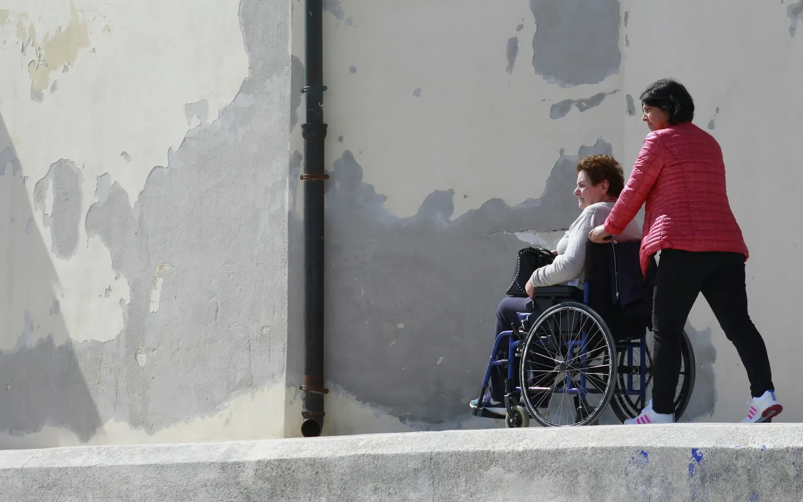 Femme qui pousse une personne âgée en fauteuil roulant le long d’un mur, trajet vers le véhicule d’un circuit de transport PMR