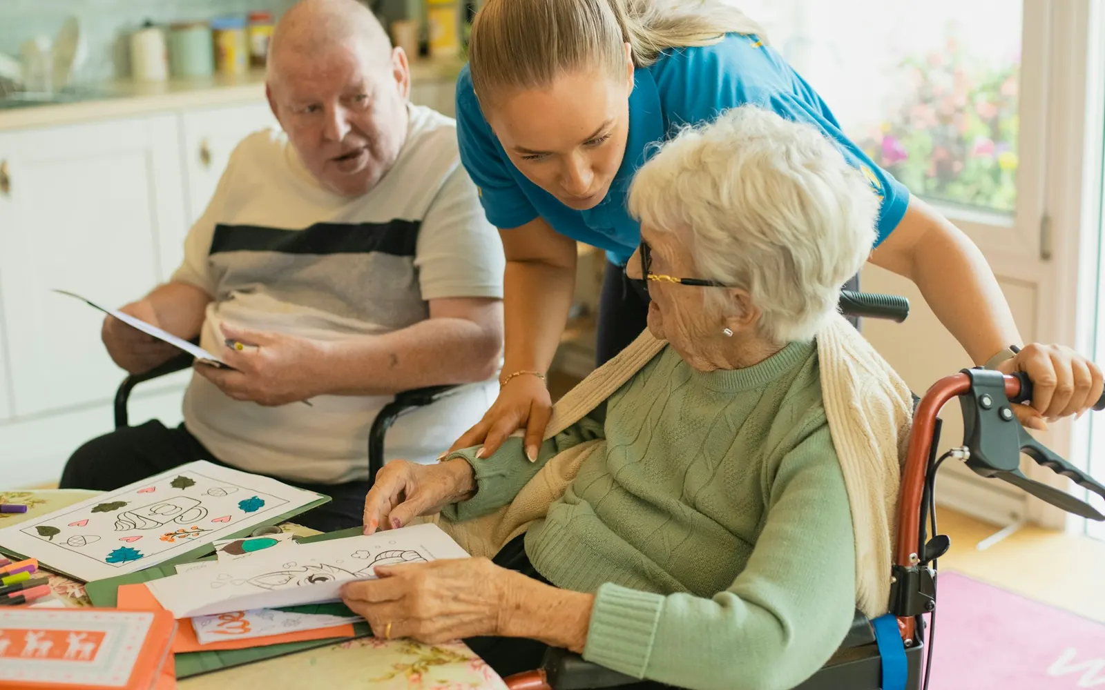 Aide-soignante avec des résidents âgés dans la salle d’activités d’un établissement, dont un en fauteuil roulant, avant un transport adapté