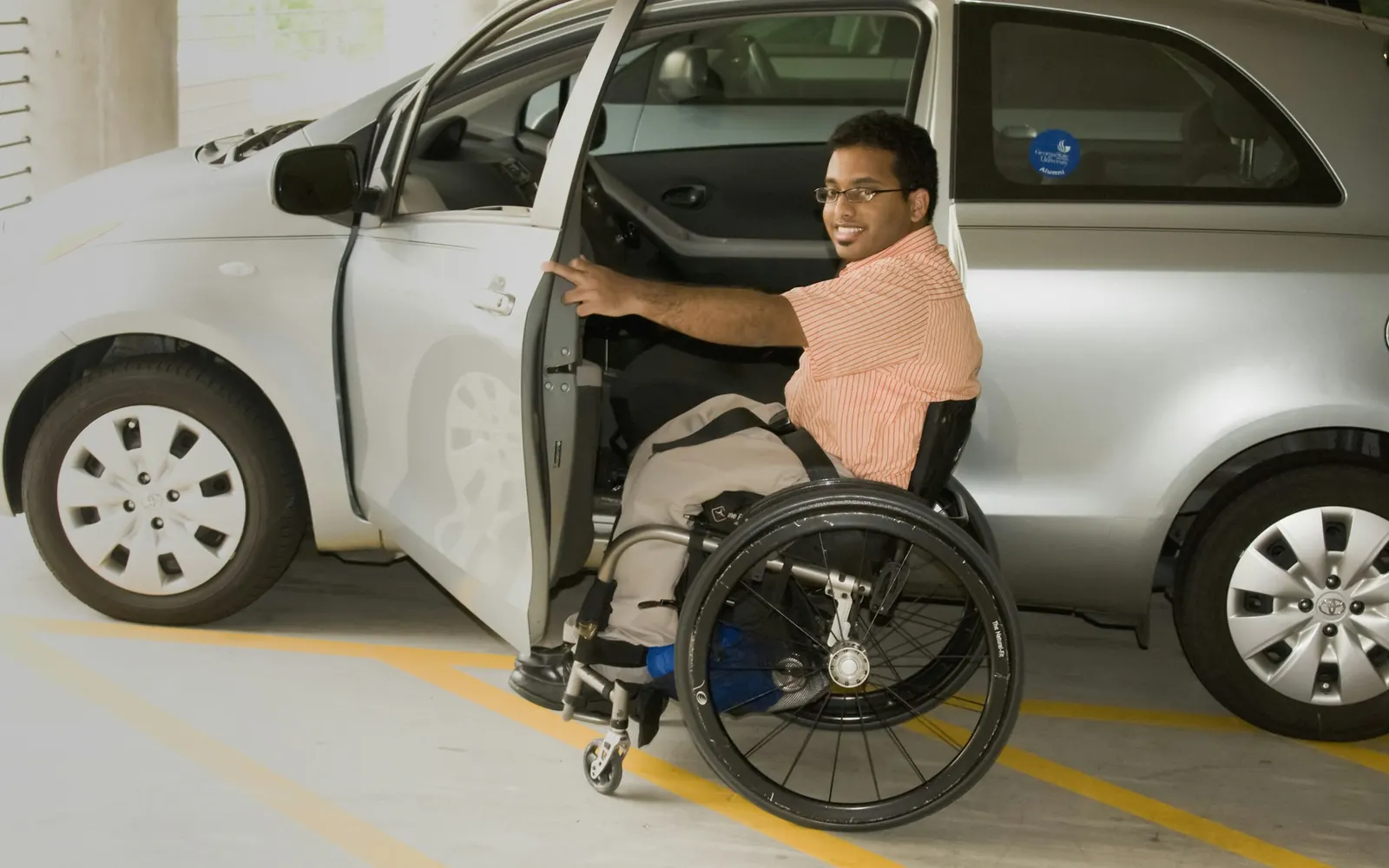 Homme en fauteuil roulant qui se transfère sur le siège d’une voiture grise, portière ouverte, sur un parking